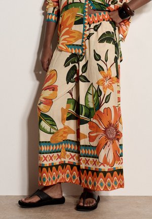 Hose mit weitem Bein und großem orange-grünem Blumenmuster, kombiniert mit passendem gemustertem Oberteil und schwarzen Riemchensandalen.