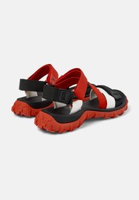 Černé a červené sandály s robustní červenou podrážkou, nastavitelnými popruhy a kombinací elastických a textilních materiálů. Mají texturovaný design.