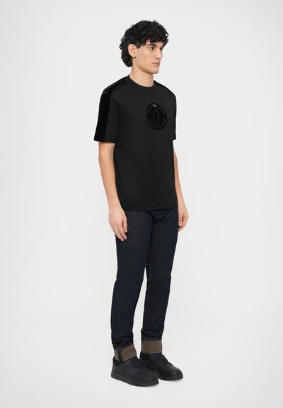 Juoda trumpomis rankovėmis marškinėliai su apvalia aksomine dėmė krūtinėje ir aksominiais akcentais ant pečių, derinami su tamsiais džinsais ir juodais sportbačiais.