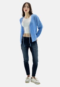 Helleblaue Strickjacke mit lockerer Passform, kombiniert mit einem weißen Crop-Top, dunklen Skinny-Jeans und weißen Sneakers mit grauen Akzenten.