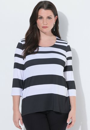 Žena s dlouhými tmavými vlasy, oblečená v černo-bílém horizontálně pruhovaném tričku s dlouhým rukávem a černých kalhotách na jednobarevném pozadí.