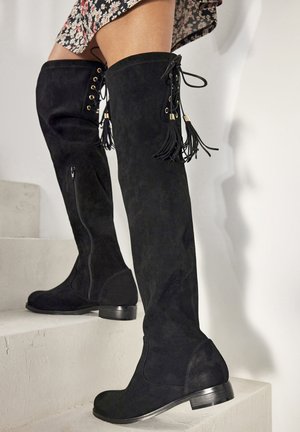 Bottes noires en suède au-dessus du genou avec une fermeture éclair sur le côté, ornées d'œillets dorés et de pompons noués le long de l'arrière. Talon bas et empilé.