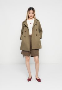 Olivgrüner doppelt knöpfbarer Trenchcoat mit braunen Knöpfen, getragen über einem weißen Hemd mit Herzdesign und braunen strukturierten Shorts. Rote Ballerinas.