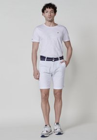 T-shirt blanche avec un petit logo, associée à un short blanc avec des poignets roulés, maintenu par une ceinture rayée bleu marine, et des baskets grises.