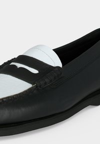 Mocassin penny en cuir noir et blanc avec couture moc toe et petit talon, incliné vers la droite sur un fond clair.
