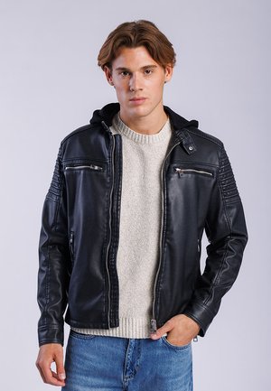 Jonge man met lichtbruin haar die een zwart leren jack draagt over een lichtgrijze trui en blauwe spijkerbroek, staand met één hand in zijn zak.