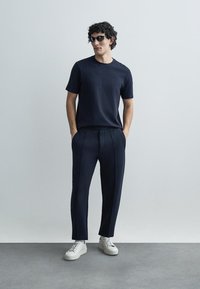 Tumši zilas teksturētas adījuma T-krekls, kopā ar piemērotiem tumši zilas krāsas biksēm ar sānu svītrām. Modelis valkā baltas kedas un tumšas saulesbrilles.