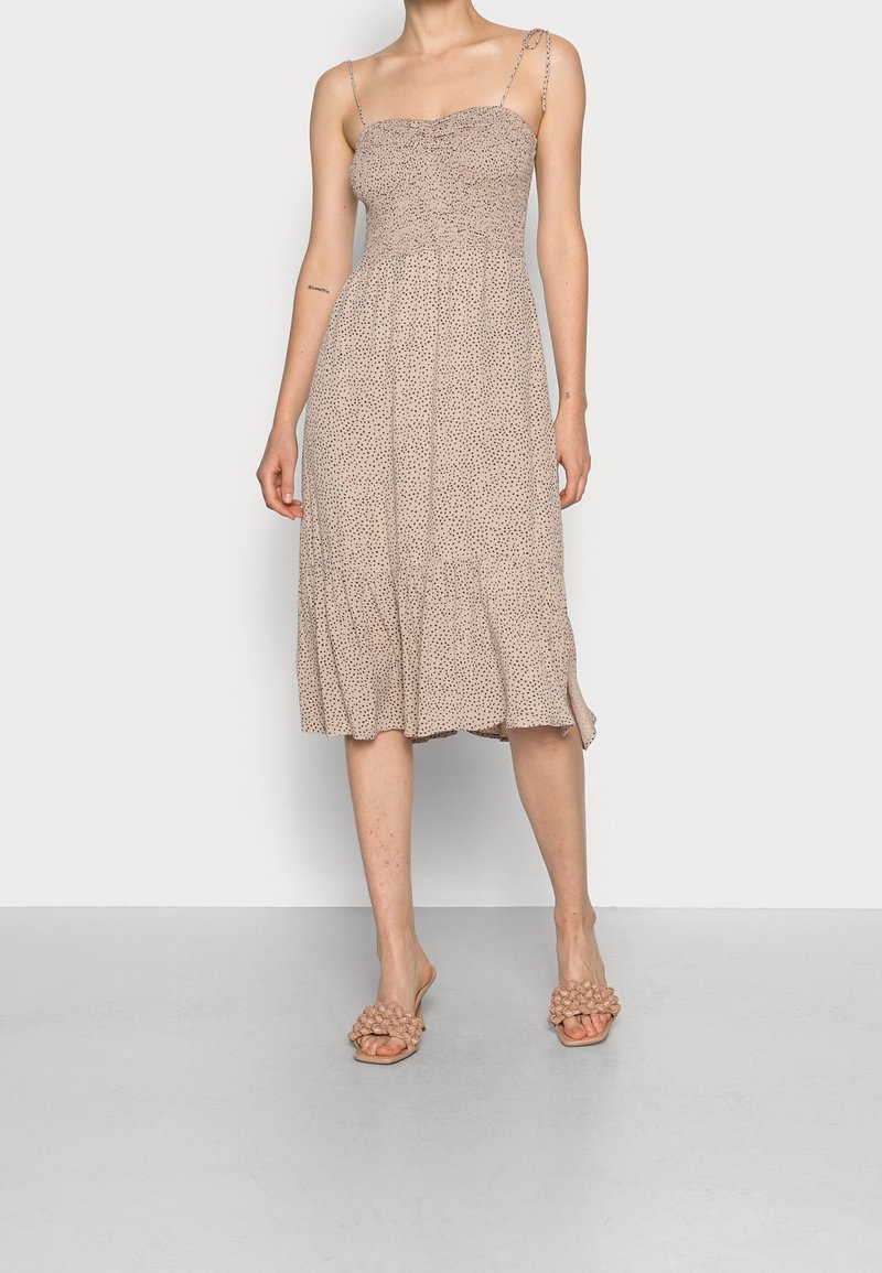 Robe mi-manche en beige avec un corsage smocké et une jupe évasée arbore un petit motif à pois. Portée avec des sandales à enfiler texturées.