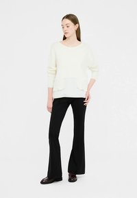 Cremefarbener Pullover mit lockerem Schnitt, Rundhalsausschnitt und vorderen Taschen, kombiniert mit schwarzen Schlaghosen und dunklen Schuhen.