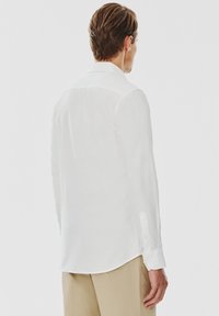 Biała longsleeve z lekkiego materiału, z klasycznym kołnierzykiem, mankietami na guziki i zaokrąglonym dołem. Tylny widok ukazuje gładką teksturę.