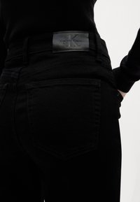 Crne traperice od denima imaju teksturiranu tkaninu, dizajn sa pet džepova i Calvin Klein kožnu oznaku blizu pojasa s reljefnim logotipom.