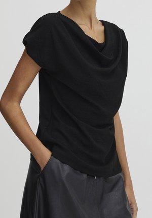 Top nero con collo a cowl realizzato in tessuto morbido, caratterizzato da maniche corte e una vestibilità rilassata. Abbinato a una gonna di pelle, ne esalta il drappeggio.