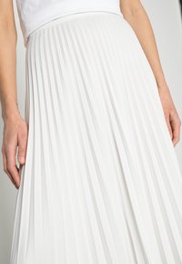 Bílá plisovaná sukně vyrobená z lehké tkaniny, s vysokým páskem a vertikálními plisami pro zvýraznění textury. Držena v ruce pro zobrazení detailu.