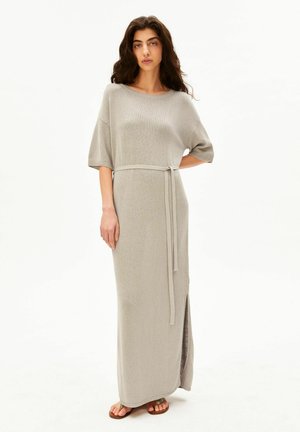 Frau mit langem dunklem Haar, die ein beiges, gestricktes Maxikleid mit kurzen Ärmeln, einem Taillengürtel, einem seitlichen Schlitz und braunen flachen Sandalen trägt.