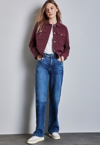 Burgunderfarbene Jacke mit Fronttaschen, weißes Hemd und hellblaue, weite Jeans. Das Outfit enthält lässige weiße Sneakers und hat eine glatte Textur.