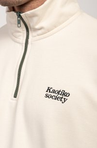 Pullover color crema con colletto alto, cerniera verde e logo ricamato "Kaotiko society" in nero sul petto. Texture liscia.