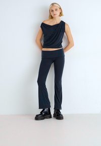 Sötétkék, aszimmetrikus dekoltázsú, drapált blúz, hozzá illő sötétkék, testhez simuló, harangszárú nadrággal párosítva. A modellen fekete, vastag talpú csizmák vannak. Fehér háttér.