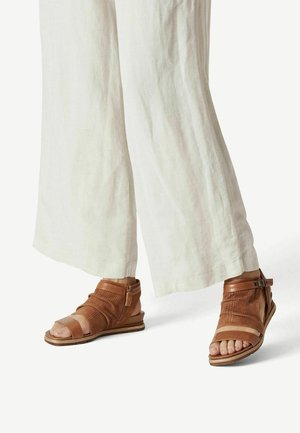 Sandales en cuir marron avec des découpes, une large bride sur le pied et une fermeture éclair à l'arrière. Portées avec un pantalon ample en lin de couleur claire.