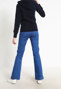 Personne de dos, portant un sweat à capuche noir, un jean évasé bleu et des baskets blanches, sur un fond clair.