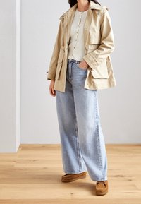Beige lättviktsjacka med huva, knäppt kofta i ljusgrått och lösa blå jeans. Bruna skor med tjock sula. Trägolv i bakgrunden.