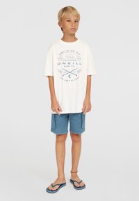 Vit bomullströja med blå tryck, lös passform. Ljusblå shorts med sidofickor, rak fåll. Svarta flip-flops med blå detaljer.