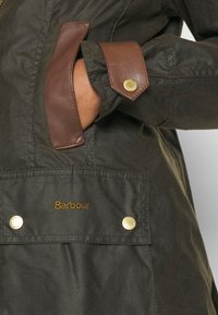 Main dans la poche d'une veste Barbour vert foncé avec des garnitures en cuir marron et des boutons-pression dorés sur la manche et le rabat de la poche.
