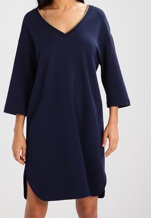 Femme portant une robe bleu marine à encolure en V avec des bordures dorées, des manches trois-quarts et des fentes latérales, posant devant un fond uni.