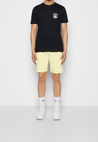 Černé bavlněné tričko s malou nášivkou, kombinované se světle žlutými šortkami a bílými vysokými teniskami. Měkká látka, volný střih.