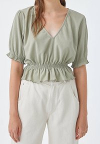Blouse vert clair texturé avec un col en V et des manches courtes à volants. Dotée d'une taille élastique et d'un ourlet style peplum.