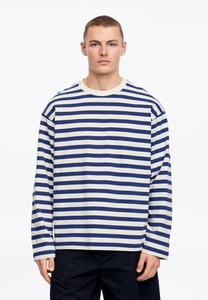 Joven con cabello corto que lleva una camisa de manga larga a rayas horizontales en azul y blanco y pantalones oscuros, mirando hacia adelante.