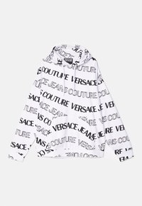Haut à capuche blanc avec un motif de texte noir répétitif portant "VERSACE" et "JEANS COUTURE." Comprend une capuche avec cordon de serrage et une poche avant.
