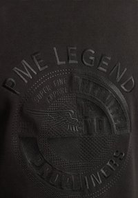 Camiseta de tela negra con texto en relieve y logo con un diseño alado. La textura es suave con un brillo sutil.