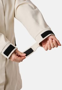 Giacca impermeabile beige chiaro con accenti neri in Velcro sui polsini, che mostra una mano che regola la cinghia in Velcro. Texture liscia e design su misura.