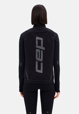 Person trägt eine schwarze ärmellose Weste mit dem "cap"-Logo in großen weißen Punkten auf dem Rücken, kombiniert mit einem schwarzen Langarmshirt und Leggings.