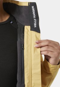 Mano sosteniendo abierta una chaqueta amarilla Helly Hansen, mostrando el forro de polar negro y un bolsillo interior con cremallera.