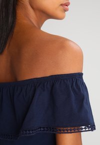 Marinblus med off-shoulder design som har en volangkrage, elastisk trim och spettdetaljer längs nederkanten. Slätt bomullstyg.