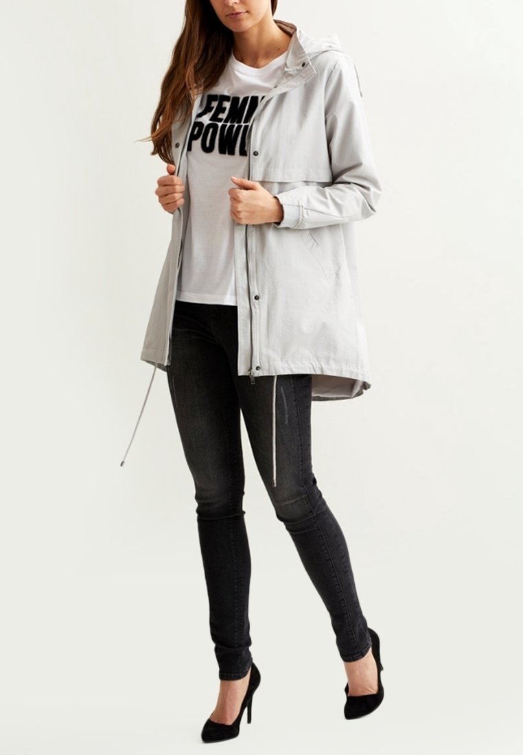 Light gray, lightweight jacket with a hood and front snap closure. Worn over a white t-shirt with bold black text, paired with dark jeans.