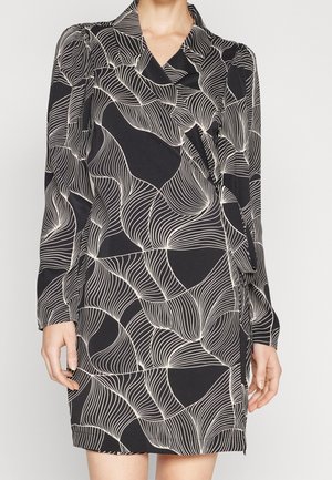 Schwarzes langärmliges Wickelkleid mit weißem abstraktem wellenförmigem Muster, weichem Kragen und leicht gepufften Schultern, getragen von einer stehenden Person.