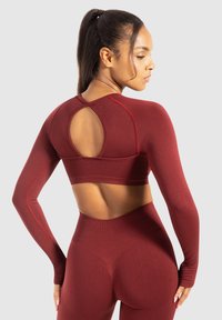 Vrouw met donker haar in een paardenstaart, gekleed in een rode lange mouw open rug crop top en bijpassende high-waisted legging, staande in zijaanzicht.