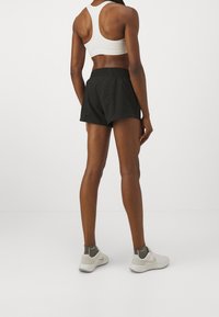 Mujer de pie, de espaldas, vistiendo un sujetador deportivo blanco, pantalones cortos deportivos negros, calcetines grises y zapatillas de running blancas, sobre un fondo blanco.