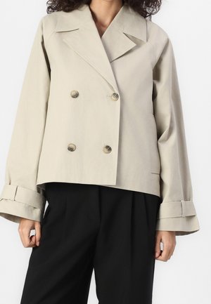 Vrouw draagt een beige, dubbelgeknoopt kort jasje met brede mouwen en zwarte, hoog opgesneden broek.