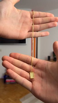 Goldkette mit einem rechteckigen Anhänger in einem perlenbesetzten Rahmen, der in einer offenen Hand präsentiert wird. Die Kette ist glatt und schlank.