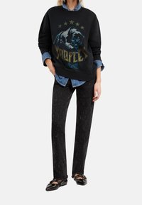 Woman wearing black sweatshirt with roaring panther graphic and gold stars, layered over blue denim shirt, black distressed jeans, and black flats.