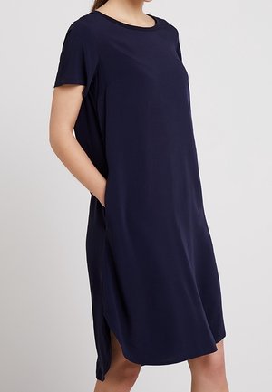 Vrouw draagt een loszittende, knielange marineblauwe jurk met korte mouwen en zijzakken, staand tegen een effen lichte achtergrond.