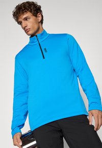 Ανοιχτό μπλε μακρυμάνικο πλεκτό με ψηλό γιακά, μαύρο φερμουάρ και υφασμάτινη υφή. Διαθέτει μια διακριτική λογότυπο στο μπροστινό μέρος.