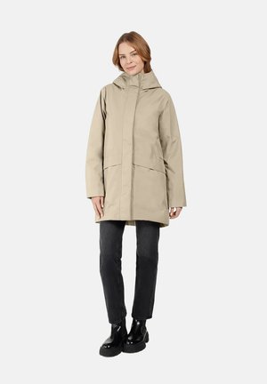 Beige waterdichte jas met een hoge kraag, lange mouwen en twee grote voorzakken. Draagt over zwarte jeans en chunky zwarte laarzen.