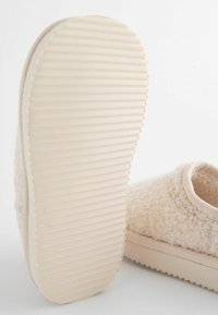 Beige toffel med en fluffig ovandel, mjukt foder och en räfflad gummisula som ger grepp och komfort.
