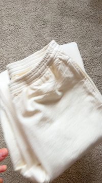 Zložene svetlo kremaste športne hlače z elastičnim pasom in teksturirano tkanino, počivajo na belem površju ob bež preprogi v ozadju.