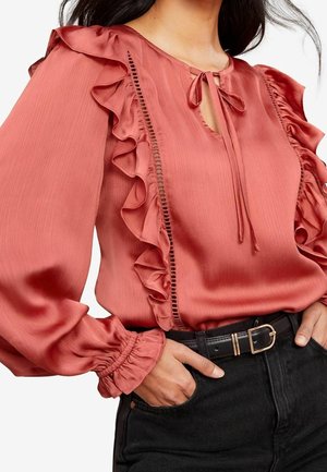 Mujer que lleva una blusa color rosa empolvado con volantes y lazos en el escote, combinada con jeans negros de talle alto y un cinturón negro.