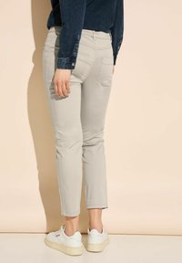 Hellgraue, schlanke Hose mit Gesäßtaschen, knöchellang, kombiniert mit weißen Sneakers. Die Denimhose hat Knopfdetails an den Ärmeln.